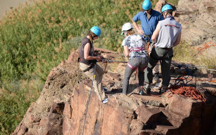 four people stand on the edge of a rock while as one person prepares to rappel down